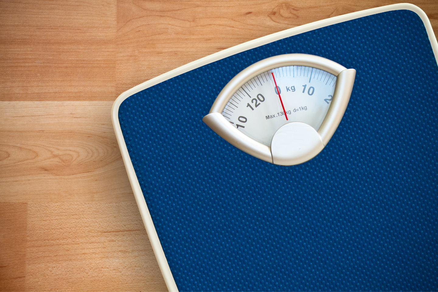 A blue analog bathroom scale with a white border rests on the wooden floor. The needle, pointing to around 118 kg, offers a reminder to treat yourself kindly as you measure your progress with care and understanding.