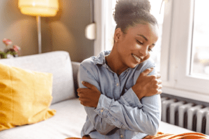 A woman with curly hair sits on a sofa, smiling and hugging herself as if to silently remind others to treat yourself kindly. Shes wearing a light blue denim shirt, with a yellow pillow behind her and warm light glowing from a lamp. Sunlight streams through the large window.
