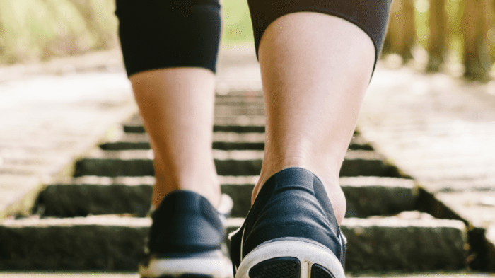 Close-up of a person wearing black sneakers and black leggings walking up outdoor stone steps, a gentle reminder to treat yourself kindly. The background is blurred with greenery, suggesting a natural setting. The image focuses on the persons legs and feet in motion.