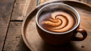 A steaming cup of bone broth sits on a wooden tray, its surface featuring a swirling, creamy pattern. The warm tones of the broth and the rustic wooden background create a cozy atmosphere reminiscent of sipping hot chocolate by a fireside.