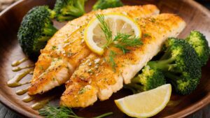 Broiled Tilapia fillets, garnished with a lemon slice and fresh dill, are served on a wooden plate alongside steamed broccoli florets and lemon wedges. The fish boasts a golden-brown crust.
