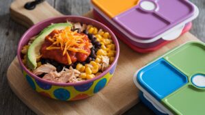 A vibrant bowl on a wooden board showcases a Chicken Burrito Bowl with shredded chicken, corn, black beans, avocado slices, chunky salsa, and shredded cheese. Nearby, two stacked lunch containers with colorful lids add a touch of convenience.