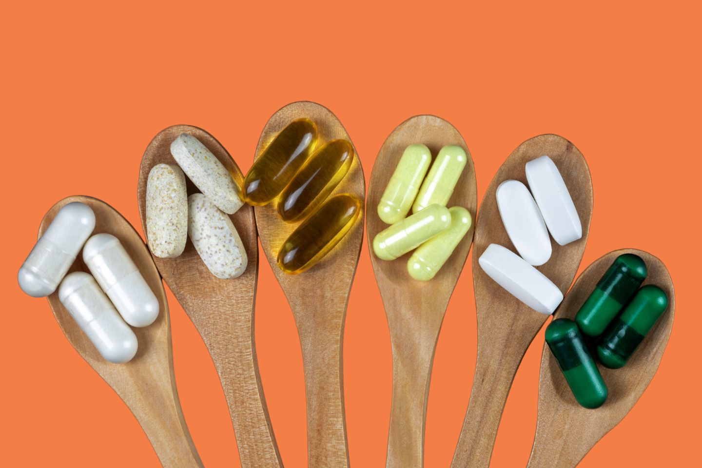 Wooden spoons arranged in a row, each holding a variety of colorful supplements and tablets, set against an orange background.
