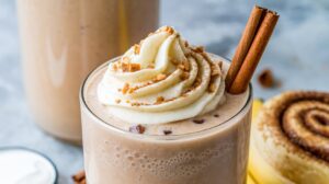 A creamy, high-protein smoothie topped with whipped cream and crushed cookies, garnished with cinnamon sticks. A cinnamon roll is partially visible in the background on a light gray surface.