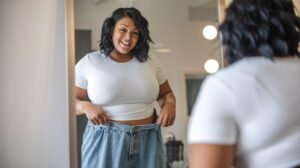 A woman who is slightly overweight is trying on a loose fitting pair of jeans and looking in the mirror smiling because the jeans are so loose.