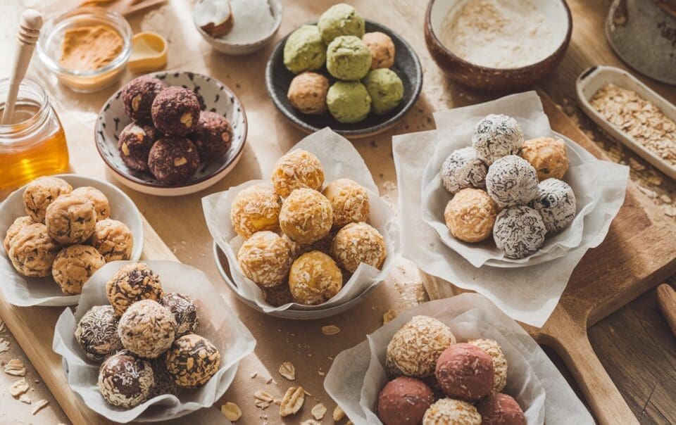 An assortment of vibrant, no-baking-required energy balls on parchment-lined bowls. Flavors like matcha, coconut, and chocolate pair with oats, honey, and cocoa on a wooden table. Snack smarter with these high-protein delights!.