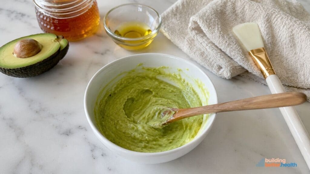A bowl of green avocado mixture for a nourishing DIY face mask sits with a wooden spoon, an avocado half, honey jar, small bowl of oil, towel, and brush on a marble surface.