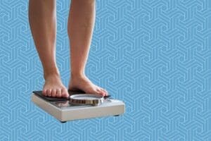 A person stands on a mechanical bathroom scale, highlighting the importance of tracking progress. Only their legs and feet are visible against a geometric blue pattern, symbolizing a journey towards weight loss.