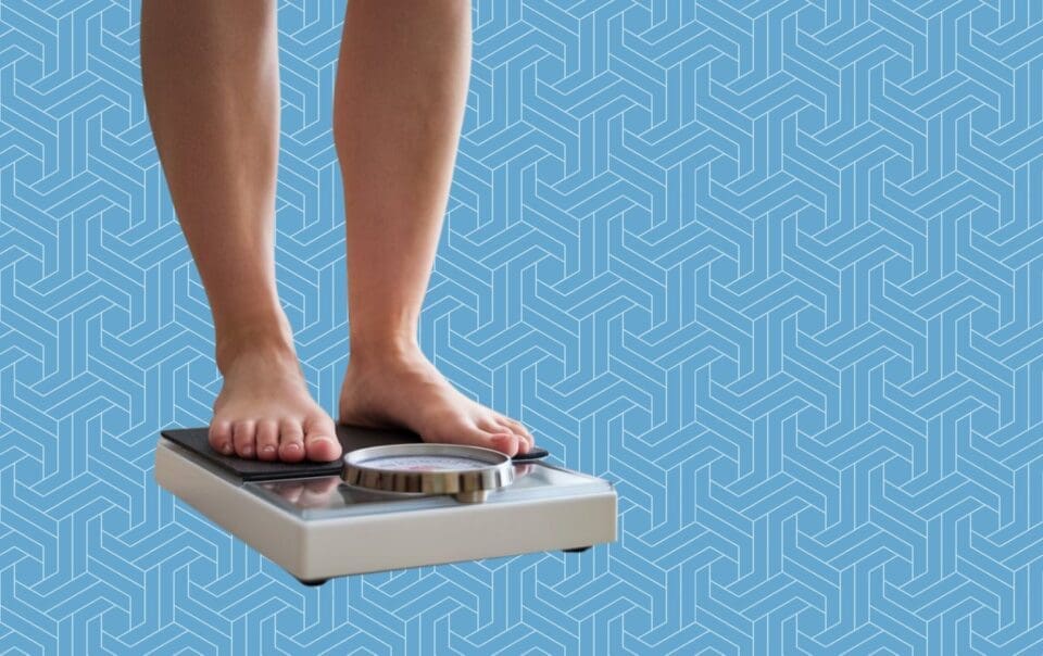 A person stands on a mechanical bathroom scale, highlighting the importance of tracking progress. Only their legs and feet are visible against a geometric blue pattern, symbolizing a journey towards weight loss.