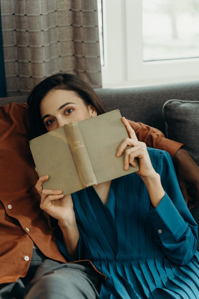 A woman in a blue dress sits on a couch, holding an open book in front of her face, with only her eyes visible over the top. She appears relaxed, seemingly lost in thought—perhaps pondering remedies for sulfur burps. A window and curtains frame the background.