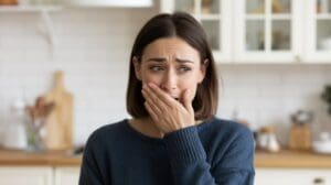 A woman with shoulder-length brown hair wearing a blue sweater covers her mouth with her hand and looks worried or anxious—possibly experiencing GLP-1 sulfur burps—while standing in a kitchen with white cabinets in the background.