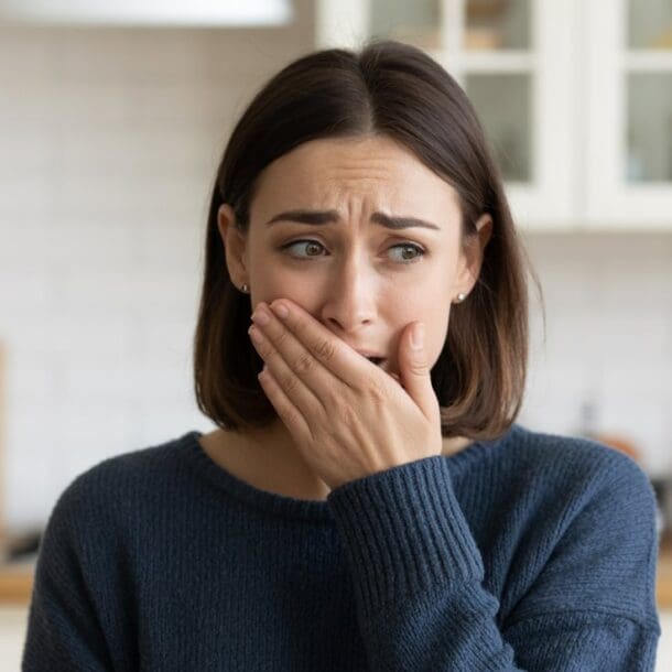 A woman with shoulder-length brown hair wearing a blue sweater covers her mouth with her hand and looks worried or anxious—possibly experiencing GLP-1 sulfur burps—while standing in a kitchen with white cabinets in the background.