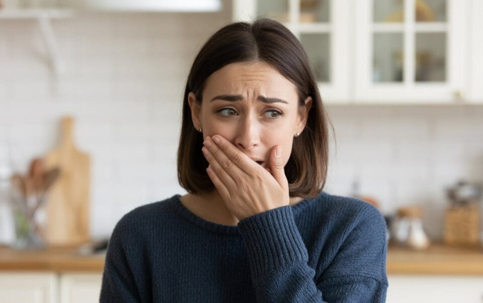 A woman with shoulder-length brown hair wearing a blue sweater covers her mouth with her hand and looks worried or anxious—possibly experiencing GLP-1 sulfur burps—while standing in a kitchen with white cabinets in the background.