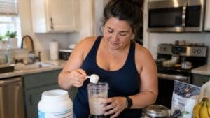 A woman in a navy tank top stands in a kitchen, smiling as she adds a scoop of supplement powder—packed with 100 grams of protein—to a blender cup. Various containers and bananas are on the counter beside her.