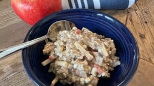 A blue bowl filled with a creamy mixture of diced apples and grains—reminiscent of Apple Cinnamon Overnight Oats—sits on a wooden table, beside a spoon and a whole red apple. A striped cloth is visible in the background.