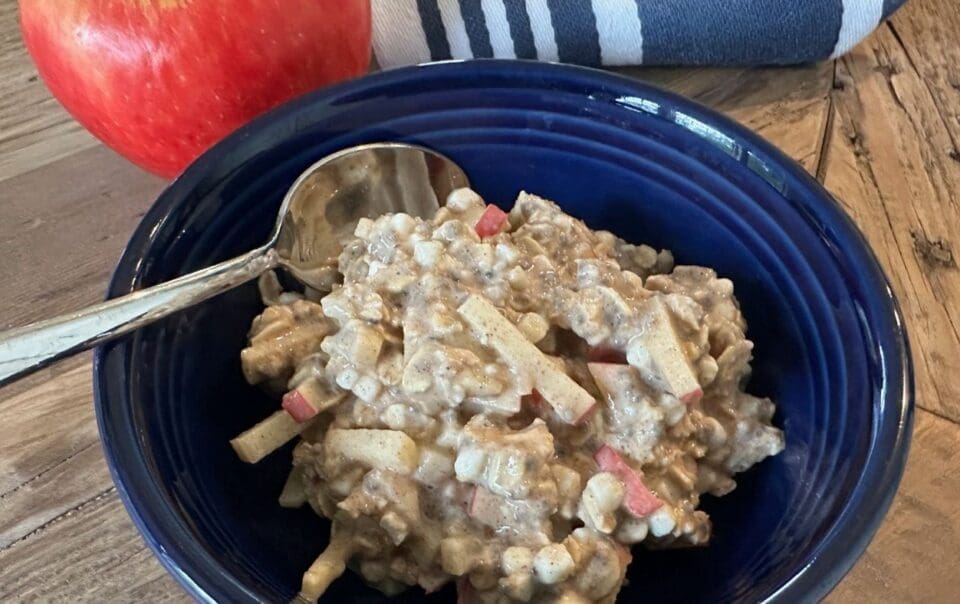 A blue bowl filled with a creamy mixture of diced apples and grains—reminiscent of Apple Cinnamon Overnight Oats—sits on a wooden table, beside a spoon and a whole red apple. A striped cloth is visible in the background.