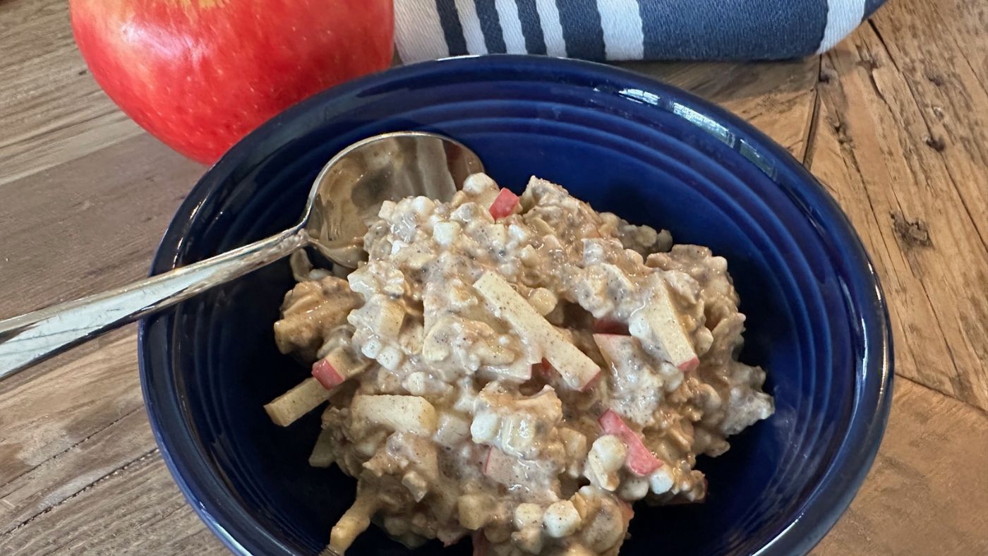 A blue bowl filled with a creamy mixture of diced apples and grains—reminiscent of Apple Cinnamon Overnight Oats—sits on a wooden table, beside a spoon and a whole red apple. A striped cloth is visible in the background.