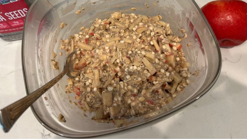 A bowl of muesli with chopped apples, seeds, and oats sits on a counter next to a fresh red apple and a container of chia seeds, evoking the wholesome flavors of apple cinnamon overnight oats. A spoon is resting in the bowl.