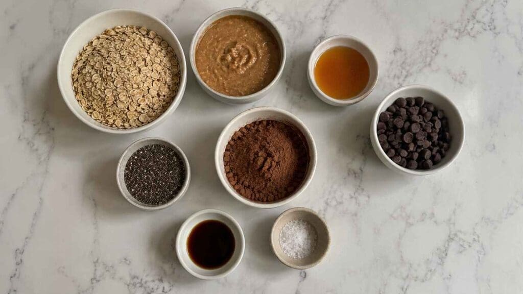 Eight bowls on a marble surface contain oats, nut butter, maple syrup, chocolate chips, chia seeds, cocoa powder, vanilla extract, and sea salt—each ingredient perfectly arranged in a circular pattern for making Chocolate Protein Balls that are high fiber thanks to the addition of nutritious chia seeds.