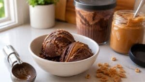 A white bowl with two scoops of Chocolate Peanut Butter protein ice cream, surrounded by a jar of peanut butter, a jar of ice cream freshly made with the Ninja Creami, a scoop, chopped peanuts, and a plant in the background.