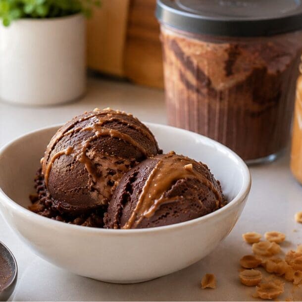 A white bowl with two scoops of Chocolate Peanut Butter protein ice cream, surrounded by a jar of peanut butter, a jar of ice cream freshly made with the Ninja Creami, a scoop, chopped peanuts, and a plant in the background.