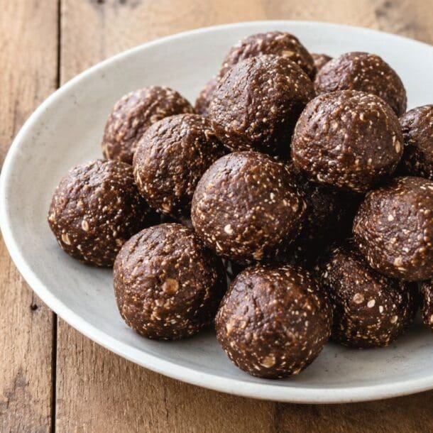 A white plate filled with round, chocolate protein balls made with oats sits on a wooden table. These high fiber energy bites are accompanied by a small bowl of chia seeds, with some seeds scattered nearby.
