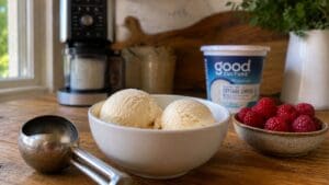 A white bowl with two scoops of ice cream, made using the Cottage Cheese Ninja Creami, sits on a wooden table next to a metal ice cream scoop, a bowl of raspberries, and a container of Good Culture cottage cheese. A coffee maker and plant are in the background.