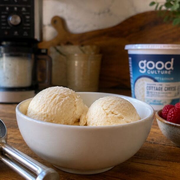 A white bowl with two scoops of ice cream, made using the Cottage Cheese Ninja Creami, sits on a wooden table next to a metal ice cream scoop, a bowl of raspberries, and a container of Good Culture cottage cheese. A coffee maker and plant are in the background.