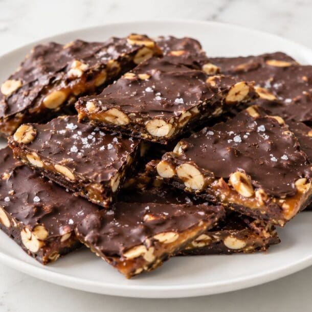 A white plate holds several pieces of chocolate bark with visible nuts and a sprinkle of sea salt, arranged neatly on a marble surface—this easy, no bake treat is the perfect vegan recipe for satisfying your sweet tooth.