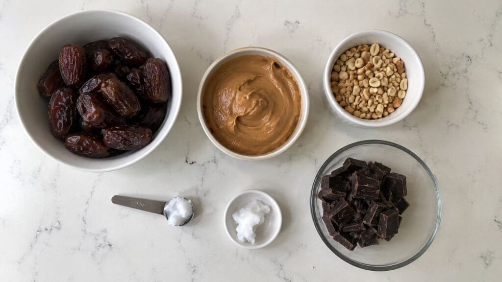 Six bowls and measuring spoons with dates, peanut butter, peanuts, chocolate chunks, coconut oil, and sea salt arranged on a white marble countertop—everything you need for this easy Vegan Recipe: No Bake Date Snickers Bars.