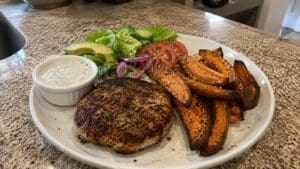 A plate featuring Easy Turkey Burgers with a grilled patty, sweet potato wedges, sliced tomato, red onion, avocado, lettuce, and a side of creamy dip sits on a granite countertop.