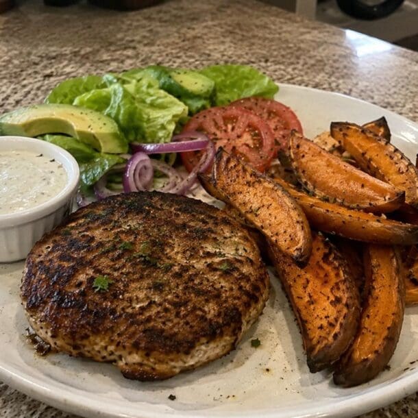 A plate featuring Easy Turkey Burgers with a grilled patty, sweet potato wedges, sliced tomato, red onion, avocado, lettuce, and a side of creamy dip sits on a granite countertop.