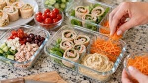 Hands preparing high protein cold lunch meal prep containers with roll-up sandwiches, shredded carrots, hummus, spinach, cherry tomatoes, cucumber, black olives, feta, and tuna on a kitchen counter. Healthy ingredients are neatly arranged.