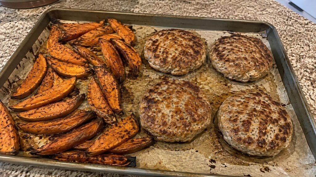 A baking sheet with four cooked Easy Turkey Burgers on the right and roasted Sweet Potato Wedges on the left, resting on a kitchen counter.