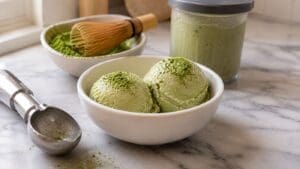 Two scoops of high protein Matcha Ice Cream in a white bowl, sprinkled with matcha powder, sit on a marble counter with an ice cream scoop, a jar of matcha, and a bamboo whisk nearby—a deliciously creamy treat made effortlessly with the Ninja Creami.