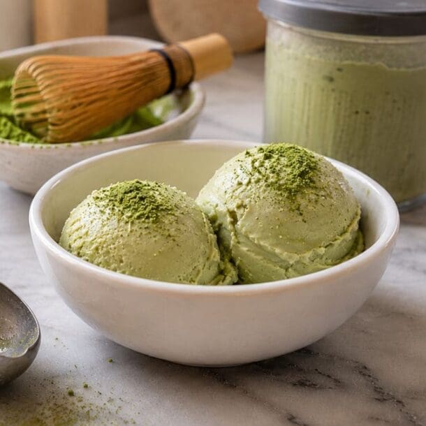 Two scoops of high protein Matcha Ice Cream in a white bowl, sprinkled with matcha powder, sit on a marble counter with an ice cream scoop, a jar of matcha, and a bamboo whisk nearby—a deliciously creamy treat made effortlessly with the Ninja Creami.