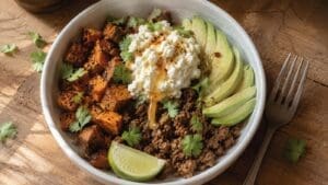 A bowl with seasoned sweet potatoes, hot honey cottage cheese, savory ground beef, sliced avocado, fresh cilantro, and a lime wedge is arranged neatly on a wooden table with a fork beside the bowl.