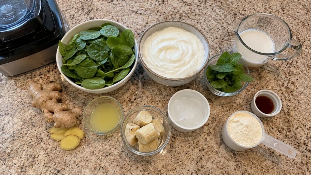 Fresh spinach, plain yogurt, milk, fresh ginger, lemon juice, banana, ice cubes, protein powder, mint leaves, and vanilla extract are arranged on a speckled countertop next to a blender—ready to be blended into a refreshing GLP-1 nausea smoothie.