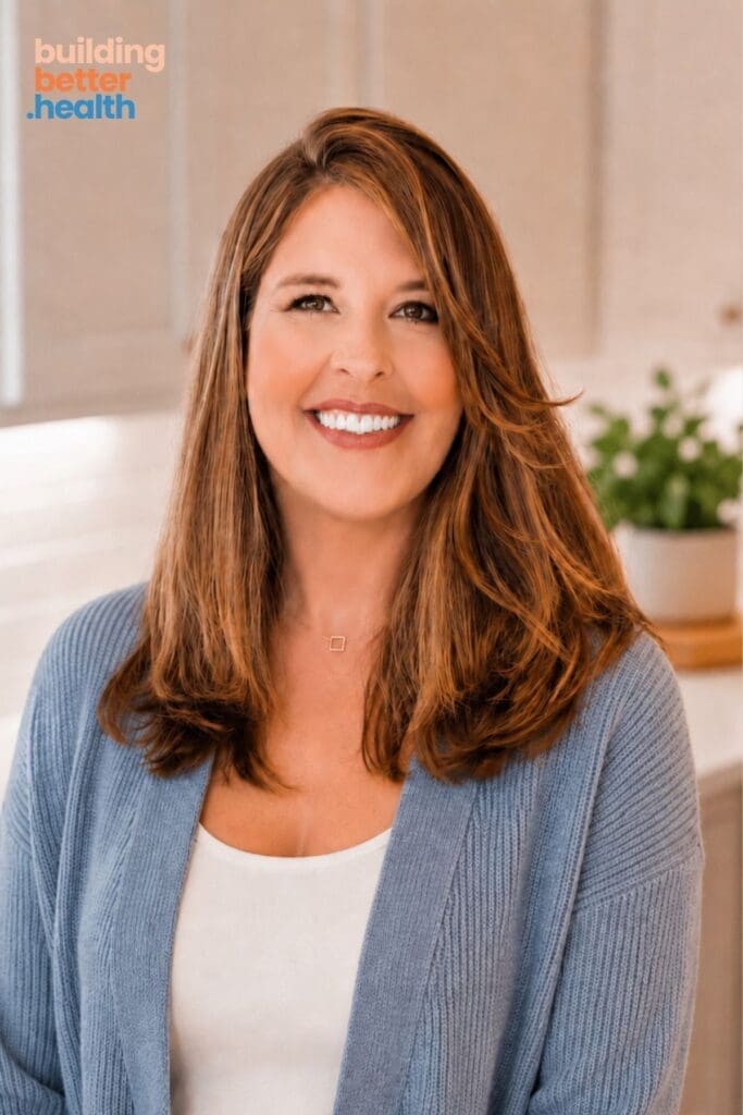 Smiling woman with long brown hair wearing a blue cardigan and white top, standing in a bright kitchen. The words about building better health are in the top left corner.