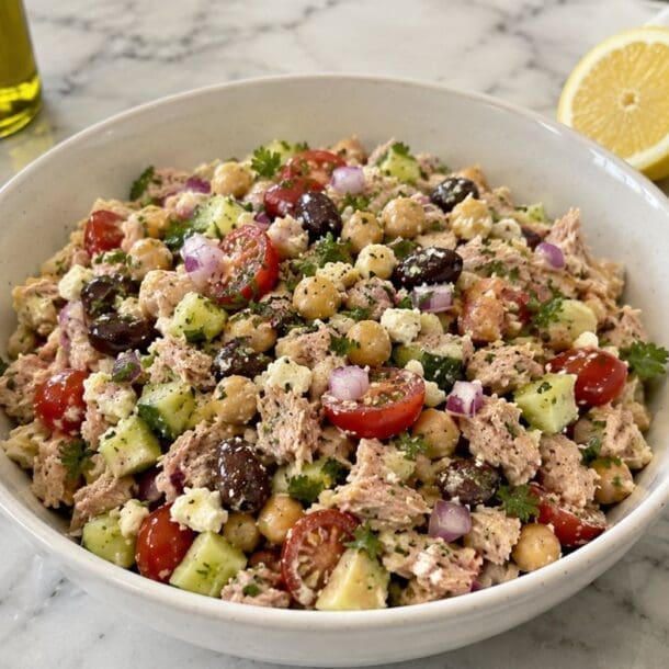 A bowl of Mediterranean Chickpea tuna salad with cherry tomatoes, cucumbers, chickpeas, olives, parsley, and red onion sits on a marble counter near a can of tuna, olive oil, a lemon half, and a wooden spoon—perfect for a fresh and easy no cook lunch.
