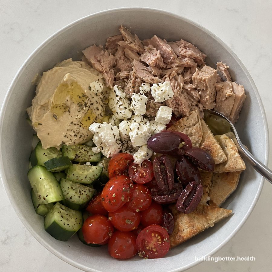 A high protein cold lunch bowl with tuna, hummus, crumbled feta, chopped cucumbers, cherry tomatoes, kalamata olives, and pita bread, all topped with fresh herbs and served with a spoon on the side.