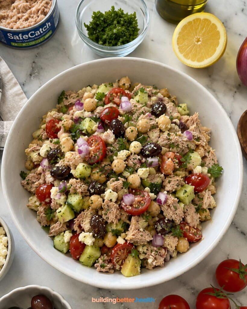 A bowl of Mediterranean Chickpea Salad, featuring a vibrant mix of tuna salad with chickpeas, cherry tomatoes, cucumber, olives, red onion, feta, and parsley. This no cook lunch recipe is completed with ingredients like lemon, herbs, and tuna arranged around the bowl on a marble surface.