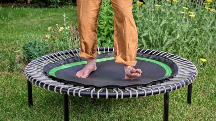 A person wearing brown pants is standing barefoot on a small round trampoline outdoors, surrounded by green grass and various yellow and red flowers.