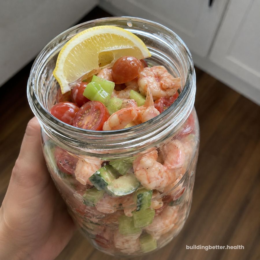 A hand holds a mason jar filled with shrimp, cucumber, celery, cherry tomatoes, and a lemon wedge on top—making for a vibrant high protein cold lunch. The jar is held above a wooden floor with white cabinets in the background.