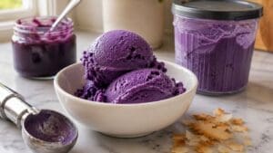 Two scoops of vibrant ube ice cream, made with the Ninja Creami for a high protein treat, sit in a white bowl next to an ice cream scoop and containers of purple spread, all displayed on a marble countertop with scattered pastry flakes.
