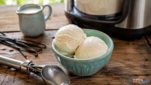 A blue bowl with two scoops of delicious vanilla protein ice cream sits on a wooden table near an ice cream maker, a metal scoop, a small pitcher, and vanilla beans—perfect for anyone craving a high protein ice cream or looking to try a new Ninja Creami recipe.