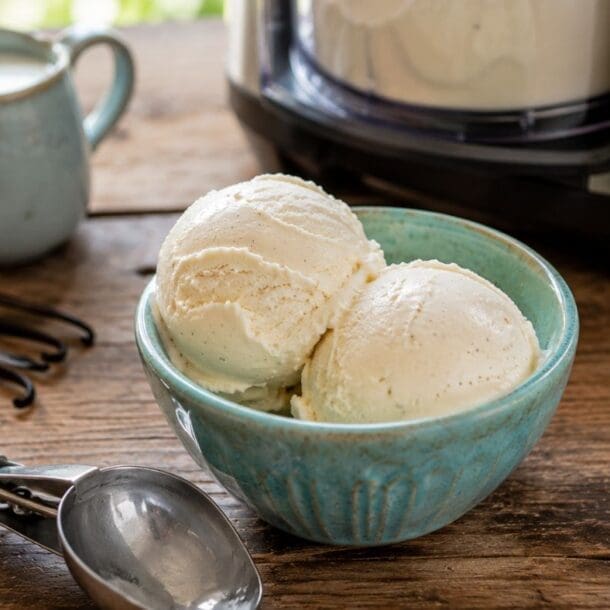 A blue bowl with two scoops of delicious vanilla protein ice cream sits on a wooden table near an ice cream maker, a metal scoop, a small pitcher, and vanilla beans—perfect for anyone craving a high protein ice cream or looking to try a new Ninja Creami recipe.