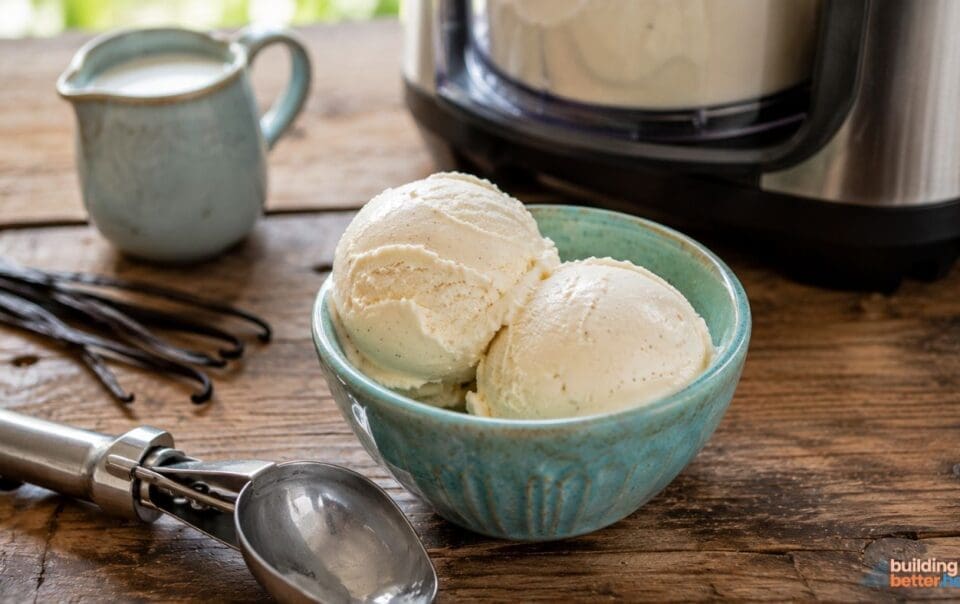 A blue bowl with two scoops of delicious vanilla protein ice cream sits on a wooden table near an ice cream maker, a metal scoop, a small pitcher, and vanilla beans—perfect for anyone craving a high protein ice cream or looking to try a new Ninja Creami recipe.