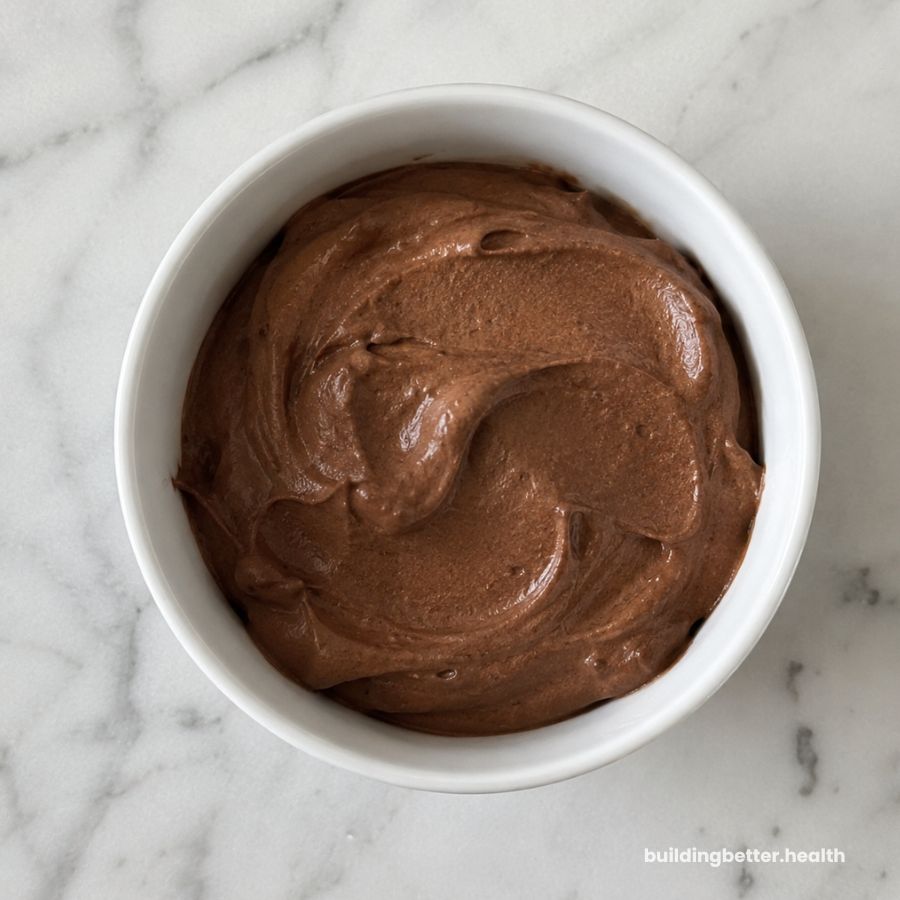 A white bowl filled with creamy, smooth chocolate mousse sits on a white marble surface. This decadent vegan dessert features a rich, swirled texture perfect for those who love dairy-free desserts.