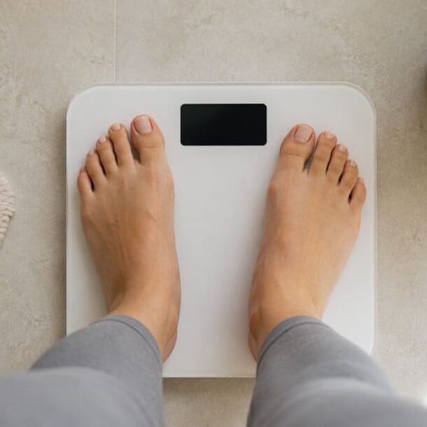 A person standing barefoot on a digital bathroom scale, with the display not showing numbers—a familiar scene for anyone experiencing a weight loss plateau, when the scale isn’t moving despite your best efforts. The floor is light-colored, and there is a green plant and a textured bath mat nearby, creating a calm and inviting atmosphere.